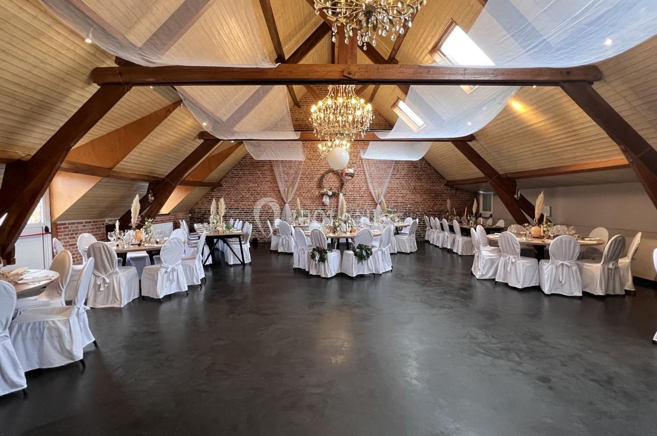 Salle de réception spacieuse avec tables décorées, chaises blanches et lustres suspendus sous un toit en bois.