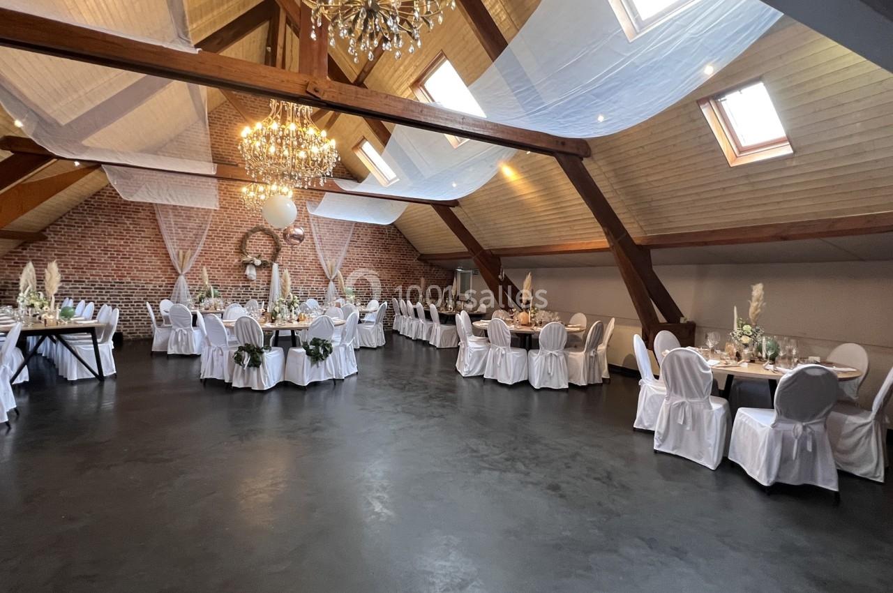 Salle de réception spacieuse avec tables décorées, chaises blanches et poutres apparentes sous un plafond en pente.