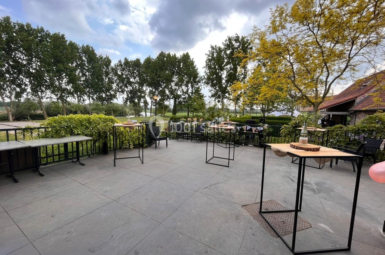 Terrasse extérieure avec tables espacées, entourée de verdure et d'arbres sous un ciel partiellement nuageux.