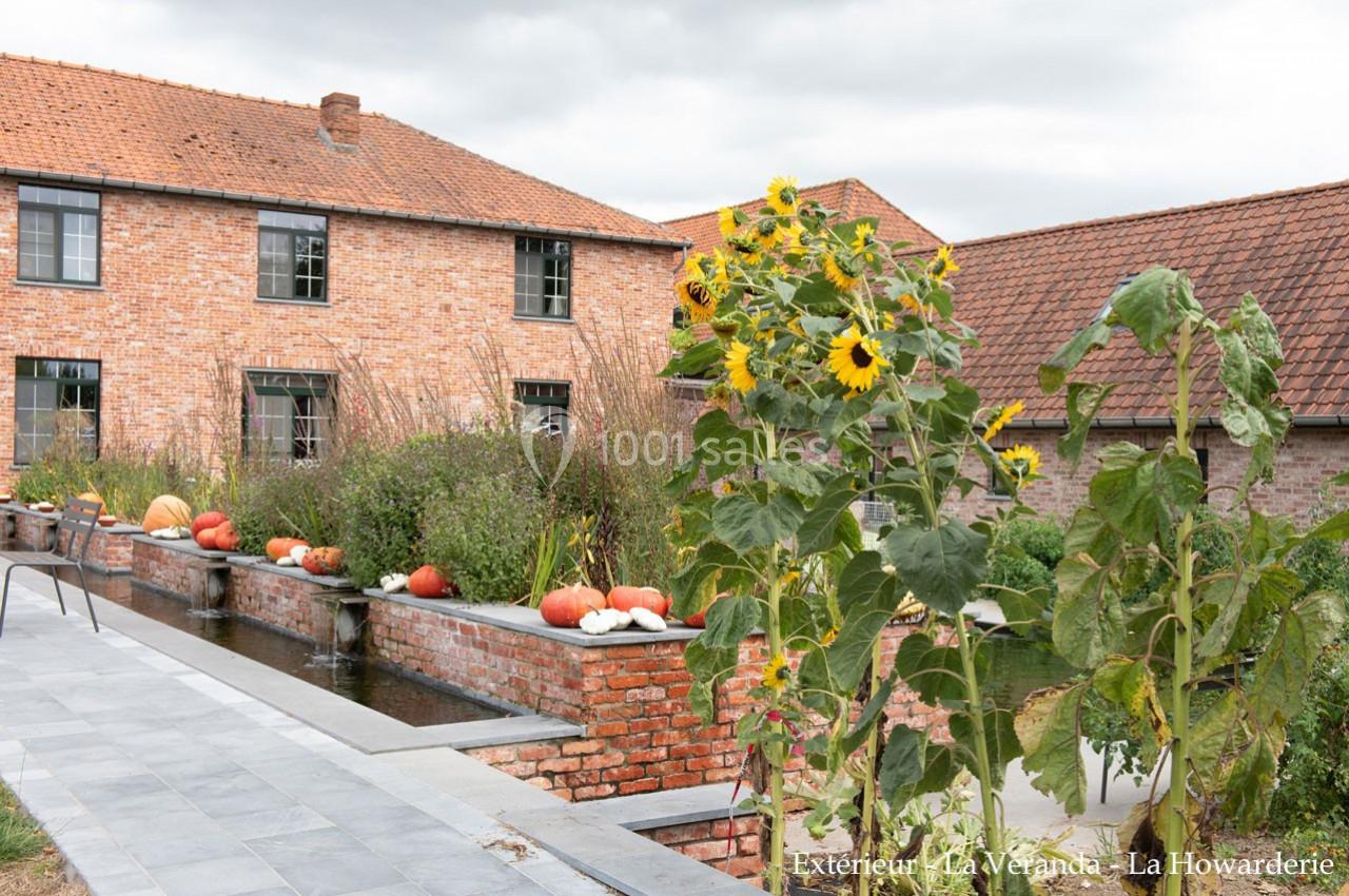 Jardin avec tournesols, citrouilles et bassin, devant des bâtiments en briques rouges sous un ciel nuageux.