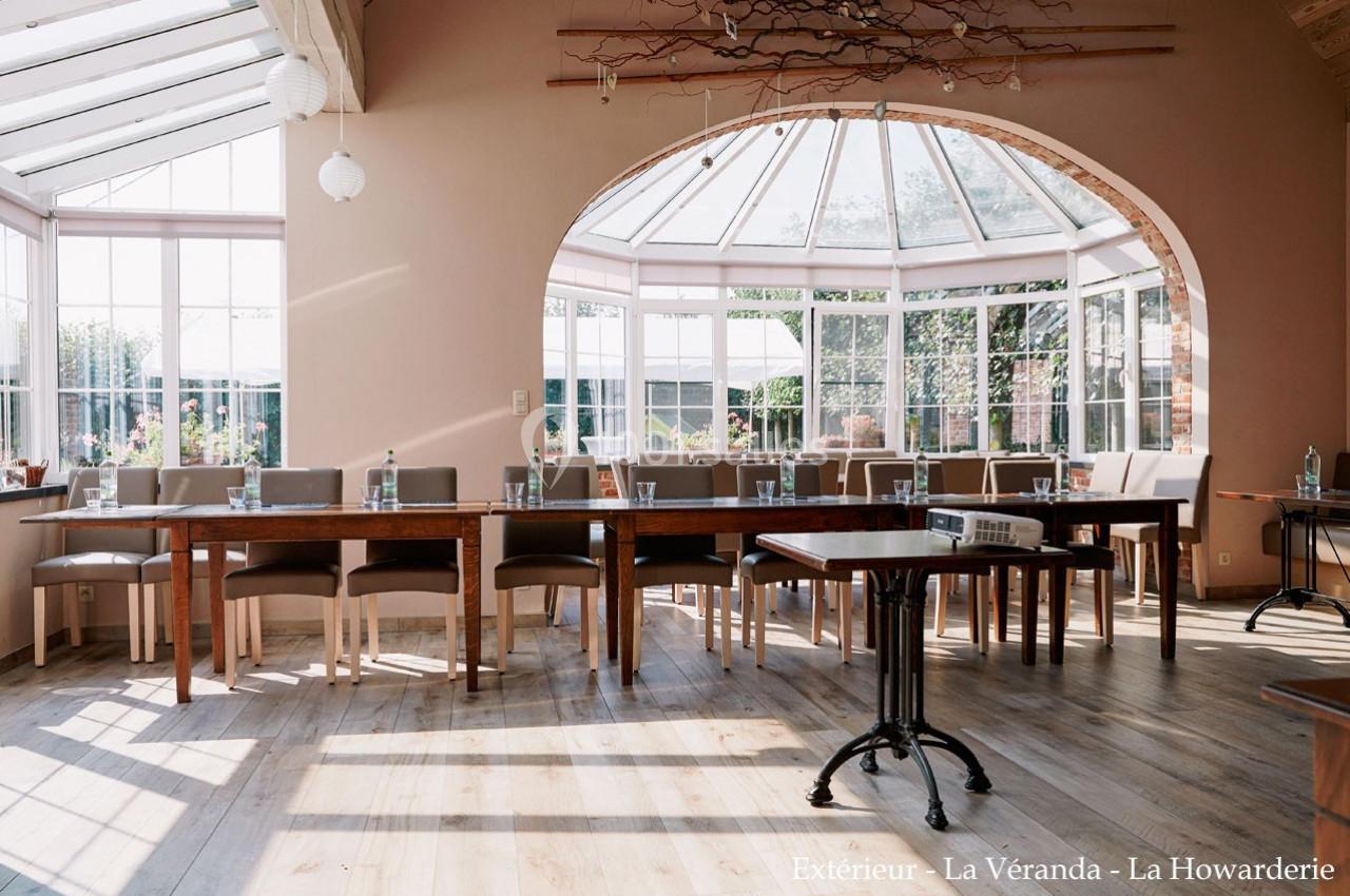 Salle lumineuse avec de grandes baies vitrées, tables en bois alignées et chaises, vue sur un jardin extérieur.