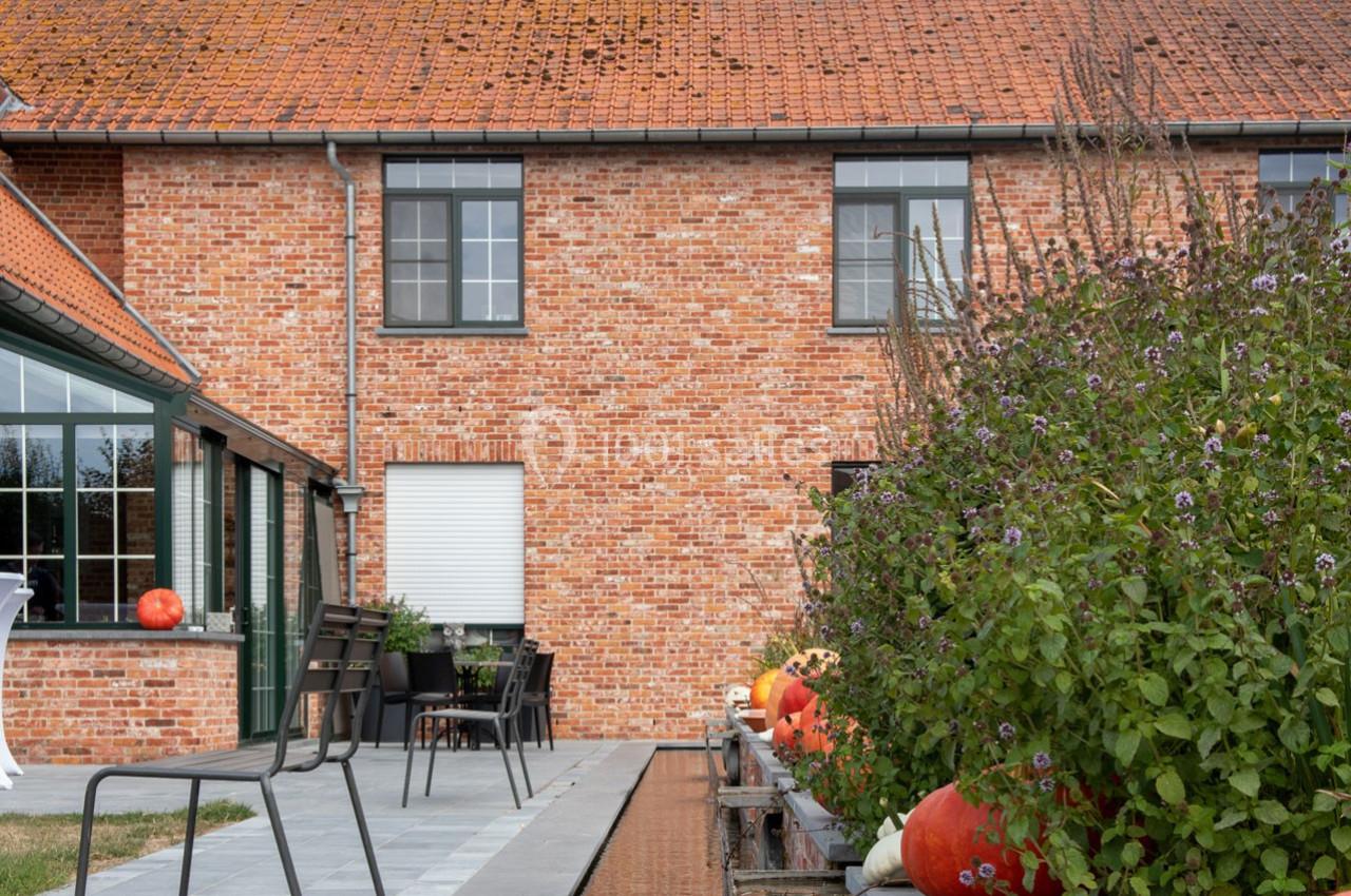 Façade en briques rouges d'une maison avec terrasse, chaises, plantes et citrouilles près d'un bassin.