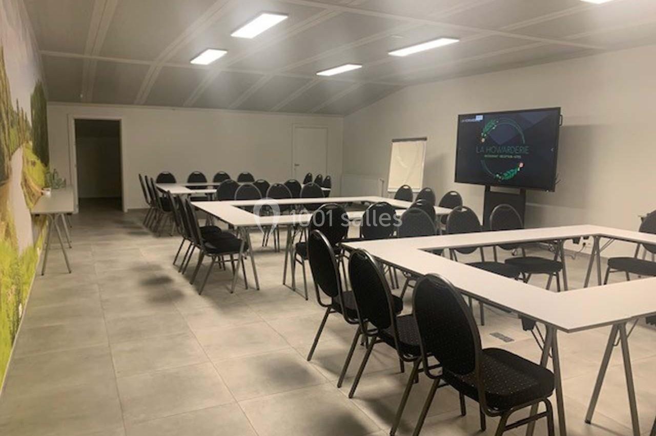 Salle de réunion lumineuse avec tables disposées en U, chaises noires, écran de présentation et tableau blanc.
