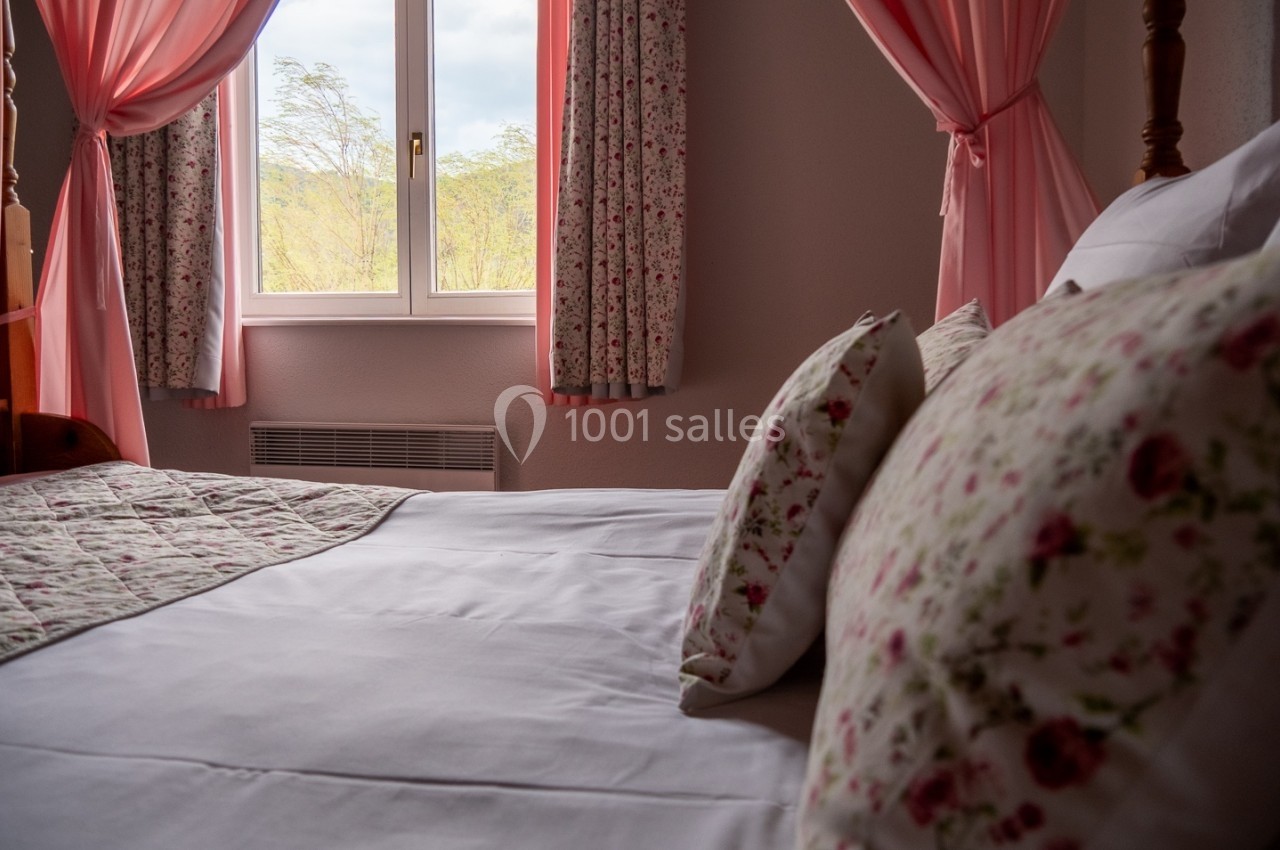 Chambre lumineuse avec lit à motifs floraux, rideaux roses et fenêtre donnant sur un paysage arboré.