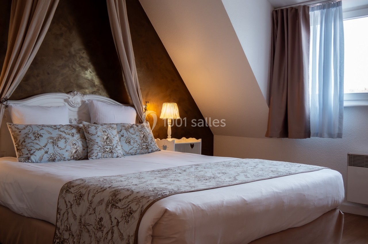 Lit double avec tête de lit blanche, draps et coussins décorés, sous un baldaquin, dans une chambre mansardée éclairée.