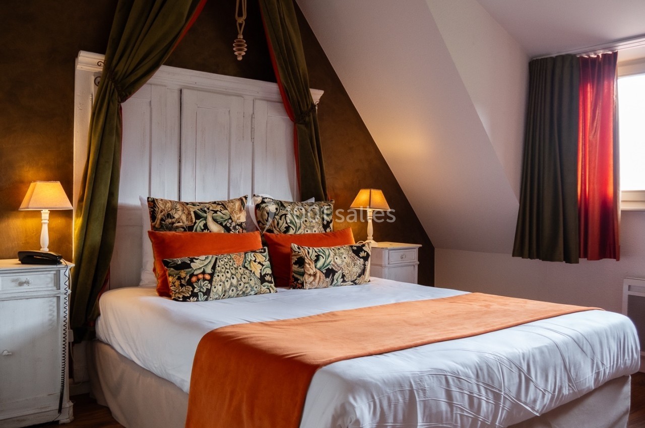 Lit double avec draps blancs, jeté de lit orange, coussins décoratifs et deux lampes de chevet dans une chambre chaleureuse.