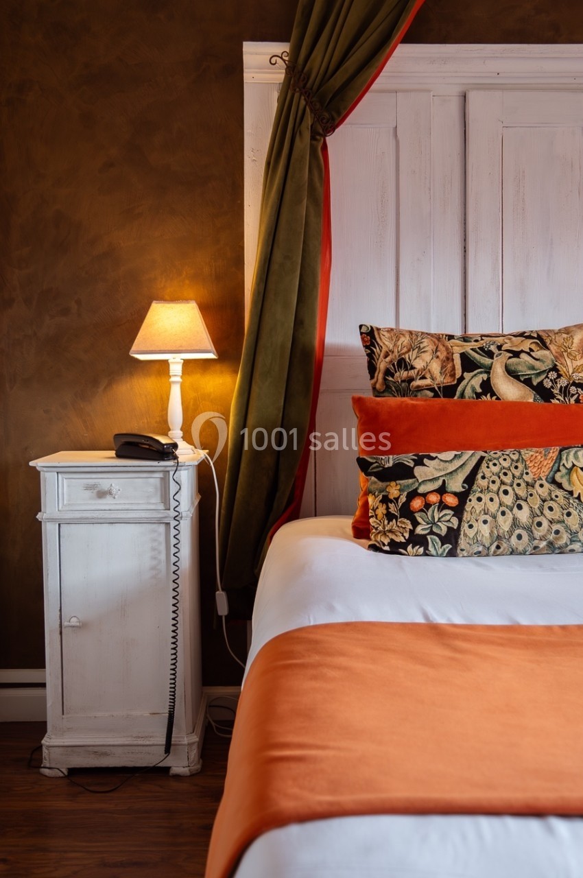 Lit avec coussins colorés, plaid orange, table de chevet blanche avec lampe allumée et rideaux verts en arrière-plan.