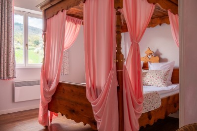 Chambre lumineuse avec lit double, murs à rayures roses, lampes de chevet et balcon donnant sur un paysage arboré.