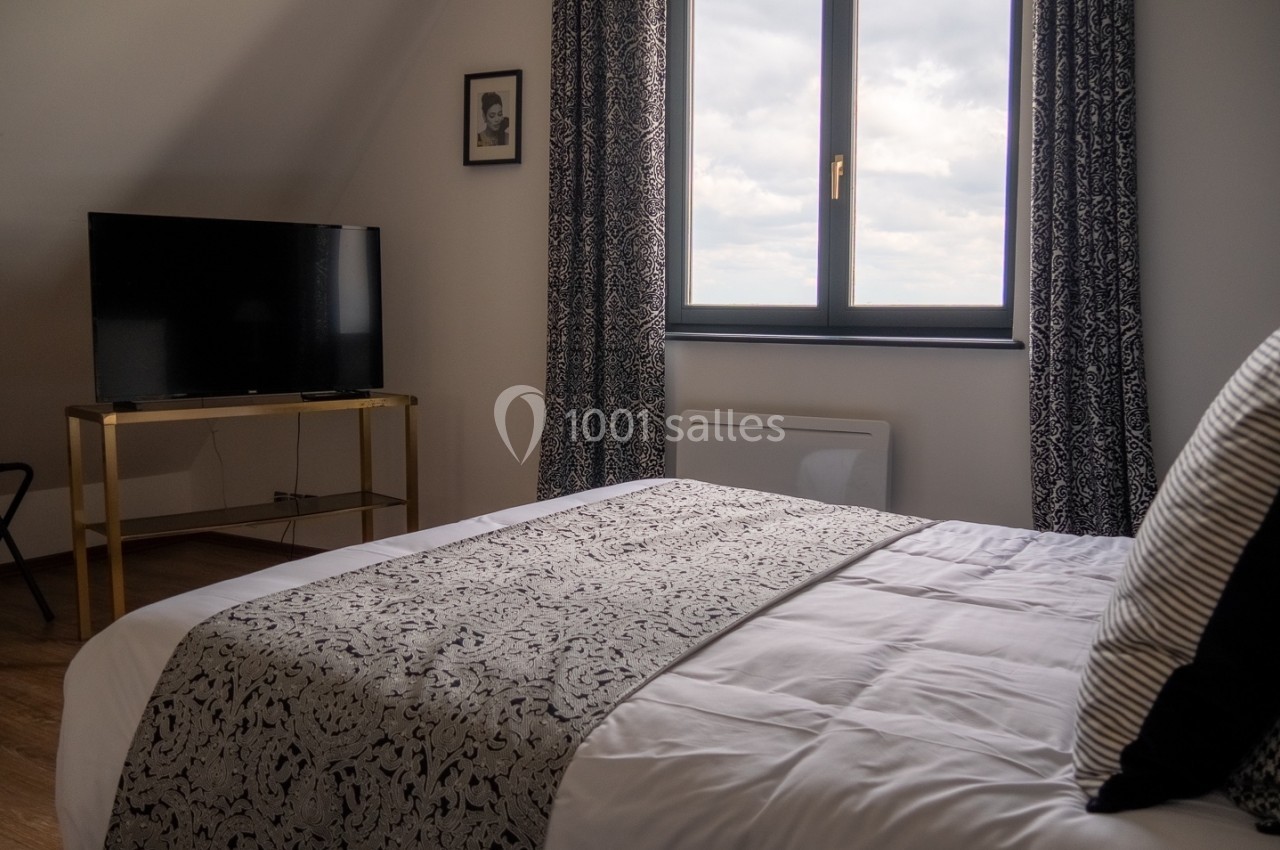 Chambre lumineuse avec lit double, télévision sur meuble doré et fenêtre donnant sur un ciel nuageux.