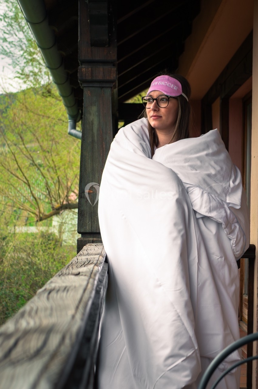 Femme enroulée dans une couverture blanche, debout sur un balcon en bois, regardant au loin un paysage verdoyant.
