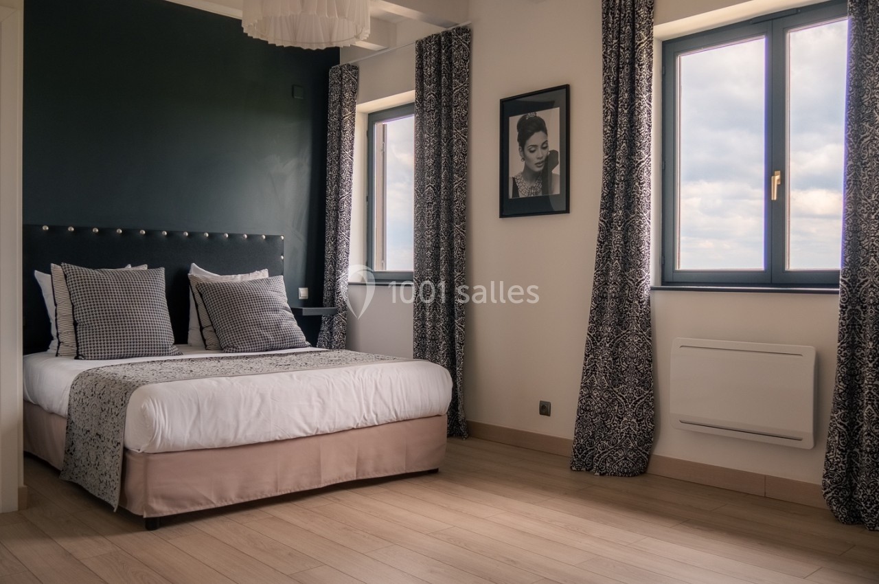 Chambre lumineuse avec lit double, murs sombres, parquet clair, deux fenêtres et un cadre décoratif.