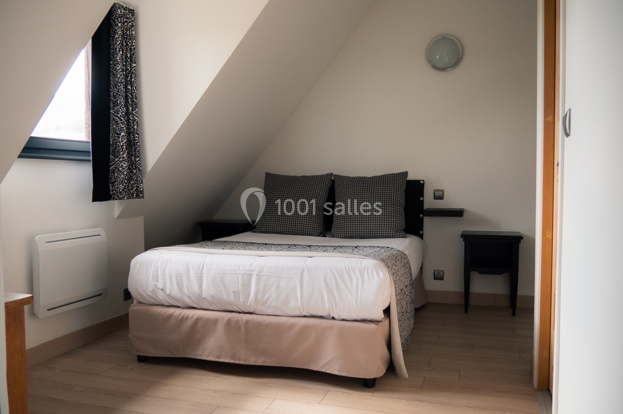 Chambre mansardée avec lit double, tables de chevet noires, parquet clair et fenêtre avec rideaux à motifs.