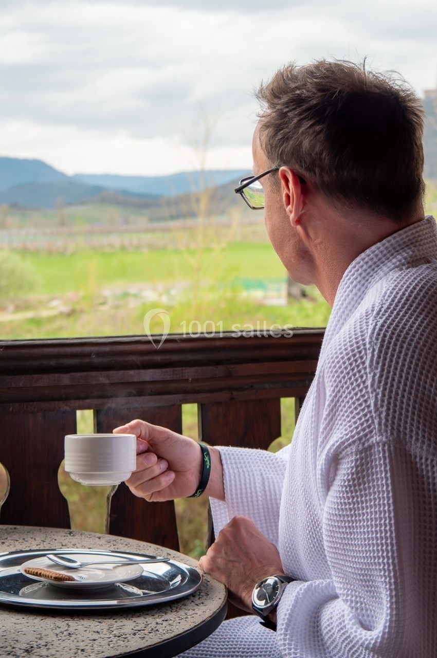 Un homme en peignoir boit une boisson chaude sur une terrasse avec vue sur une campagne vallonnée.