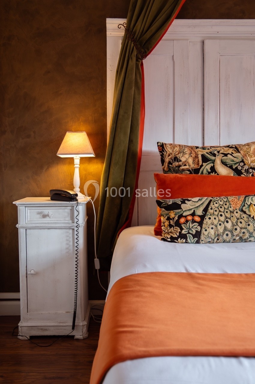 Lit avec coussins décoratifs colorés, lampe allumée sur une table de chevet blanche dans une chambre chaleureuse.