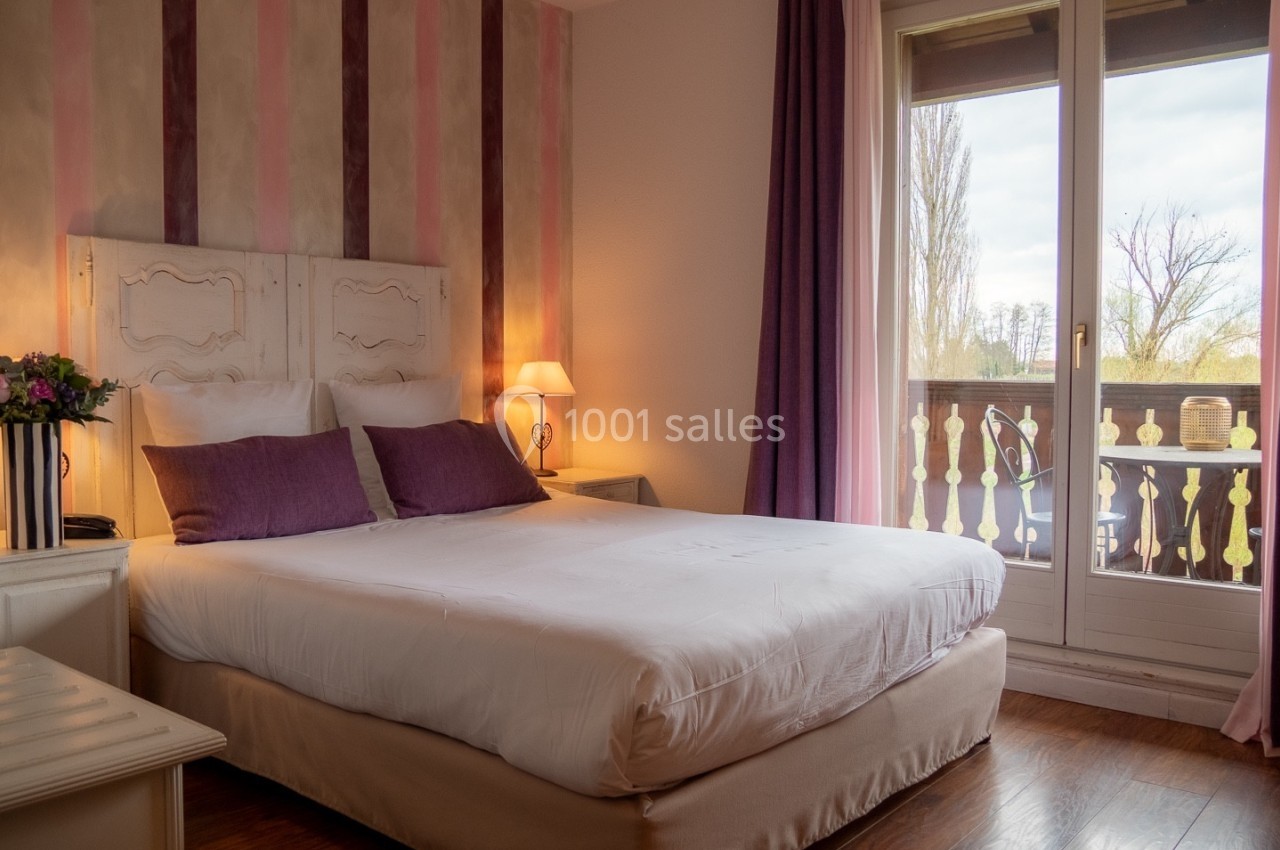Chambre lumineuse avec lit double, décoration pastel, lampes de chevet et balcon donnant sur un paysage arboré.
