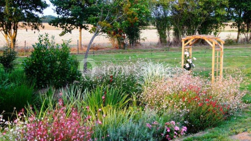 Massif de fleurs colorées entouré de verdure avec une arche en bois et des champs en arrière-plan.