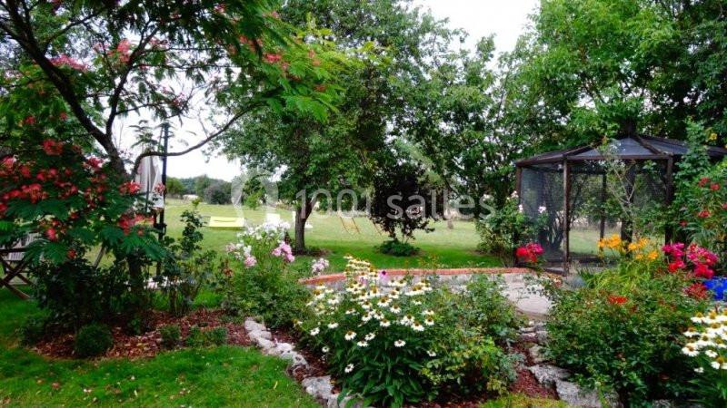 Jardin fleuri avec diverses plantes colorées, une tonnelle en verre et des arbres en arrière-plan.