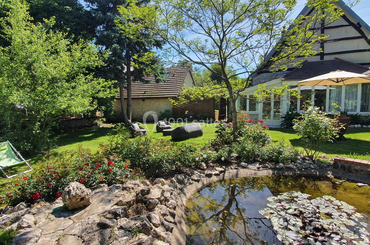 Jardin verdoyant avec un étang, des chaises longues, des fleurs et une maison à colombages en arrière-plan.