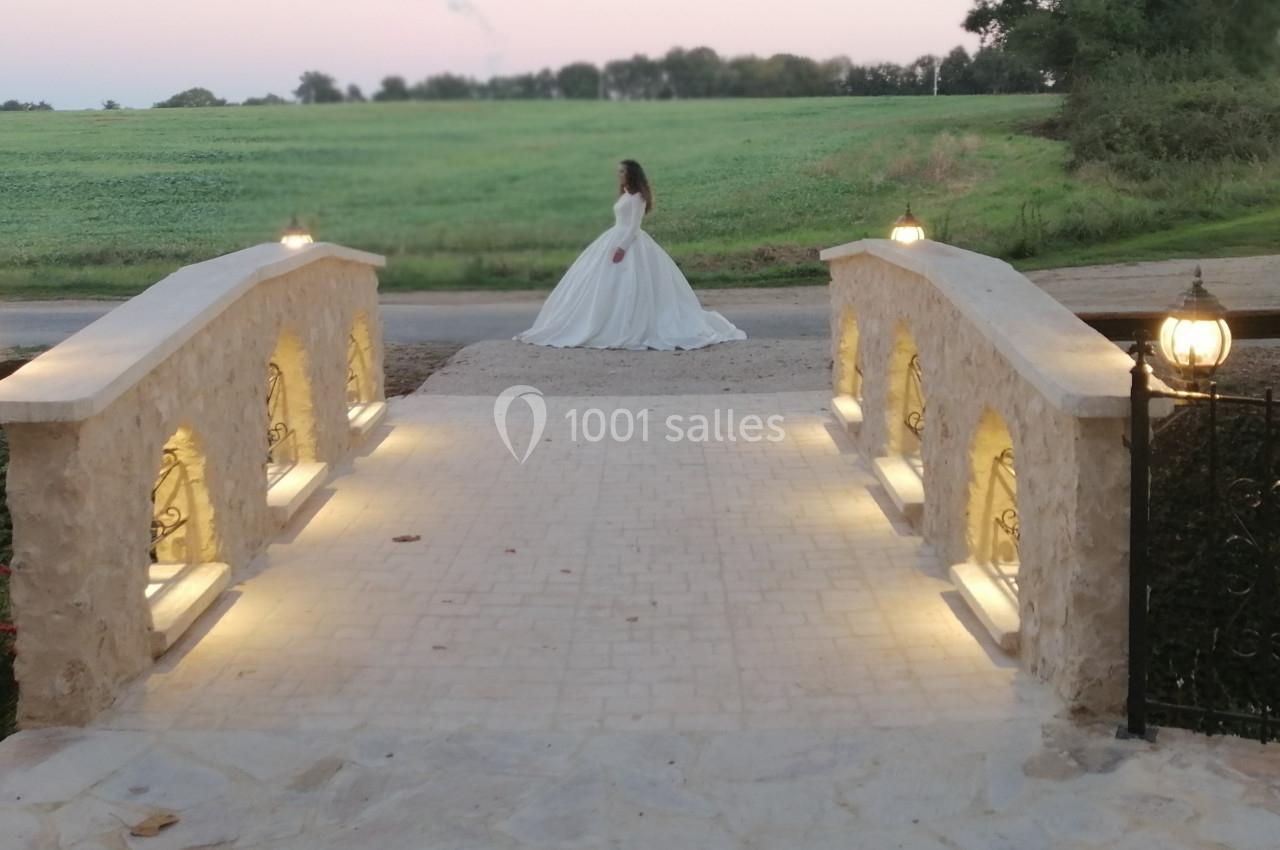 Une femme en robe blanche traverse un pont éclairé au crépuscule, avec un champ verdoyant en arrière-plan.