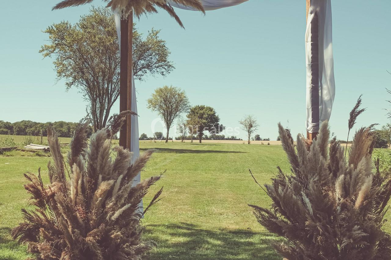 Arche en bois décorée de tissus blancs et de pampas, installée sur une pelouse avec des arbres en arrière-plan.