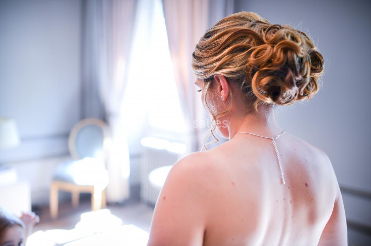 Femme de dos avec un chignon élaboré, portant un collier fin, dans une pièce lumineuse avec des rideaux et un fauteuil.