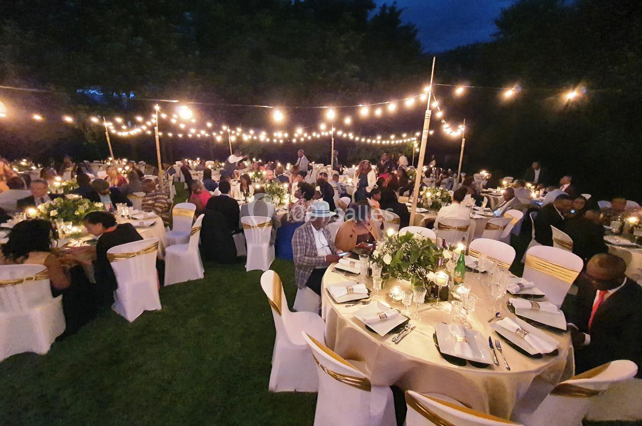 Invités assis à des tables décorées sous des guirlandes lumineuses dans un jardin en soirée.