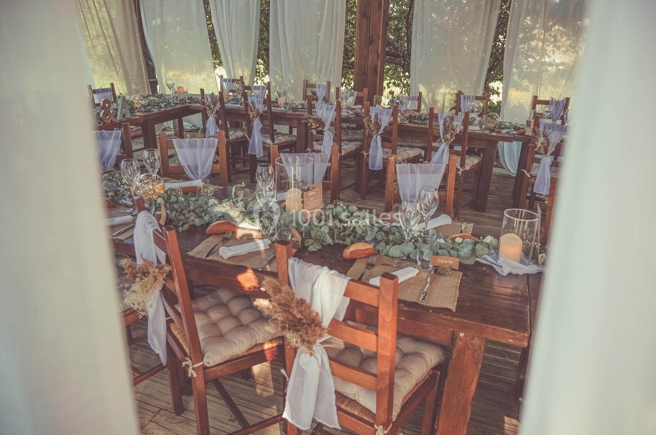 Salle de réception décorée avec des tables en bois, nappes blanches, chaises ornées et végétation en centre de table.