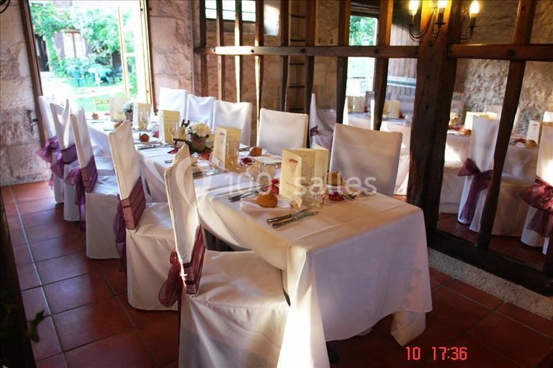Salle de réception avec tables dressées, nappes blanches, chaises décorées de rubans violets et lumière naturelle.