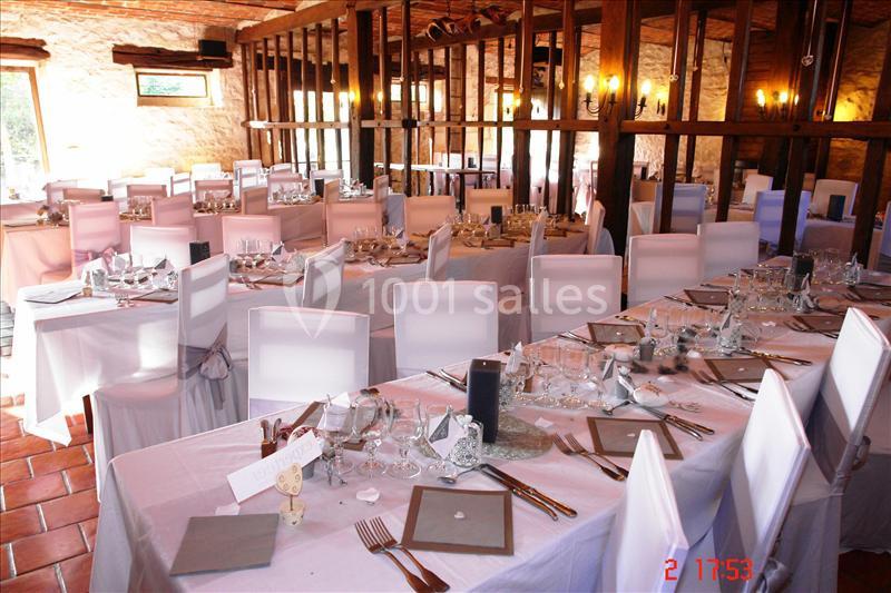 Salle de réception aménagée avec des tables dressées, nappes blanches et chaises couvertes, dans un cadre rustique.