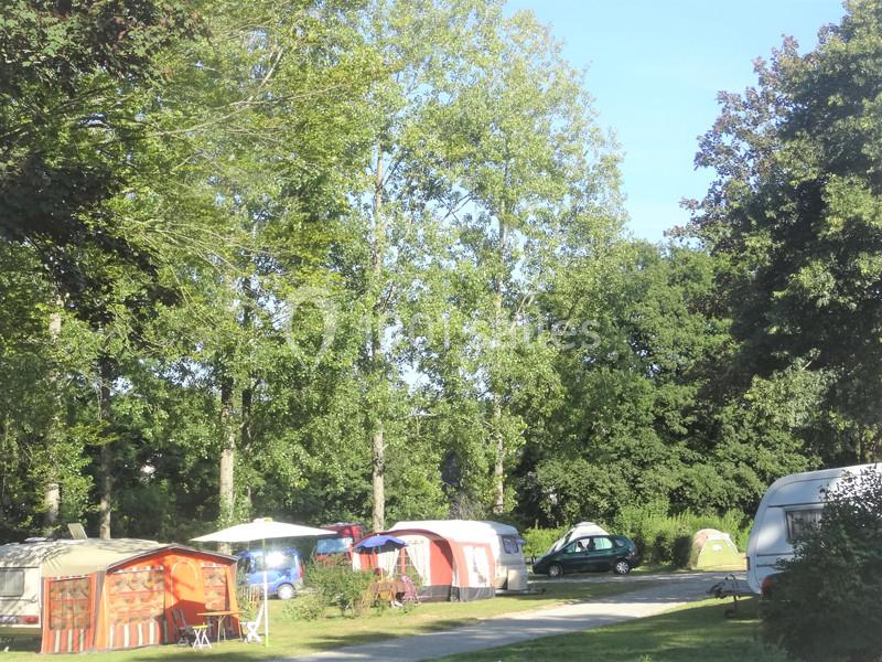Emplacements de camping avec caravanes, tentes et auvents sous des arbres dans un environnement verdoyant.