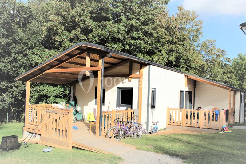 Maison en bois avec terrasse couverte, vélos et objets divers à l'extérieur, entourée de pelouse et d'arbres.