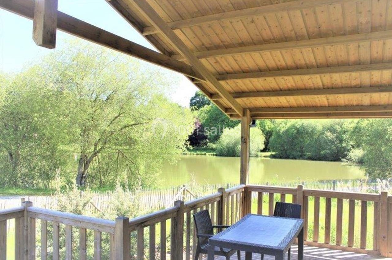 Location salle Merdrignac (Côtes-d'Armor) - Val de Landrouet #15 Terrasse en bois couverte avec table et chaises, donnant sur un étang entouré de végétation verdoyante.