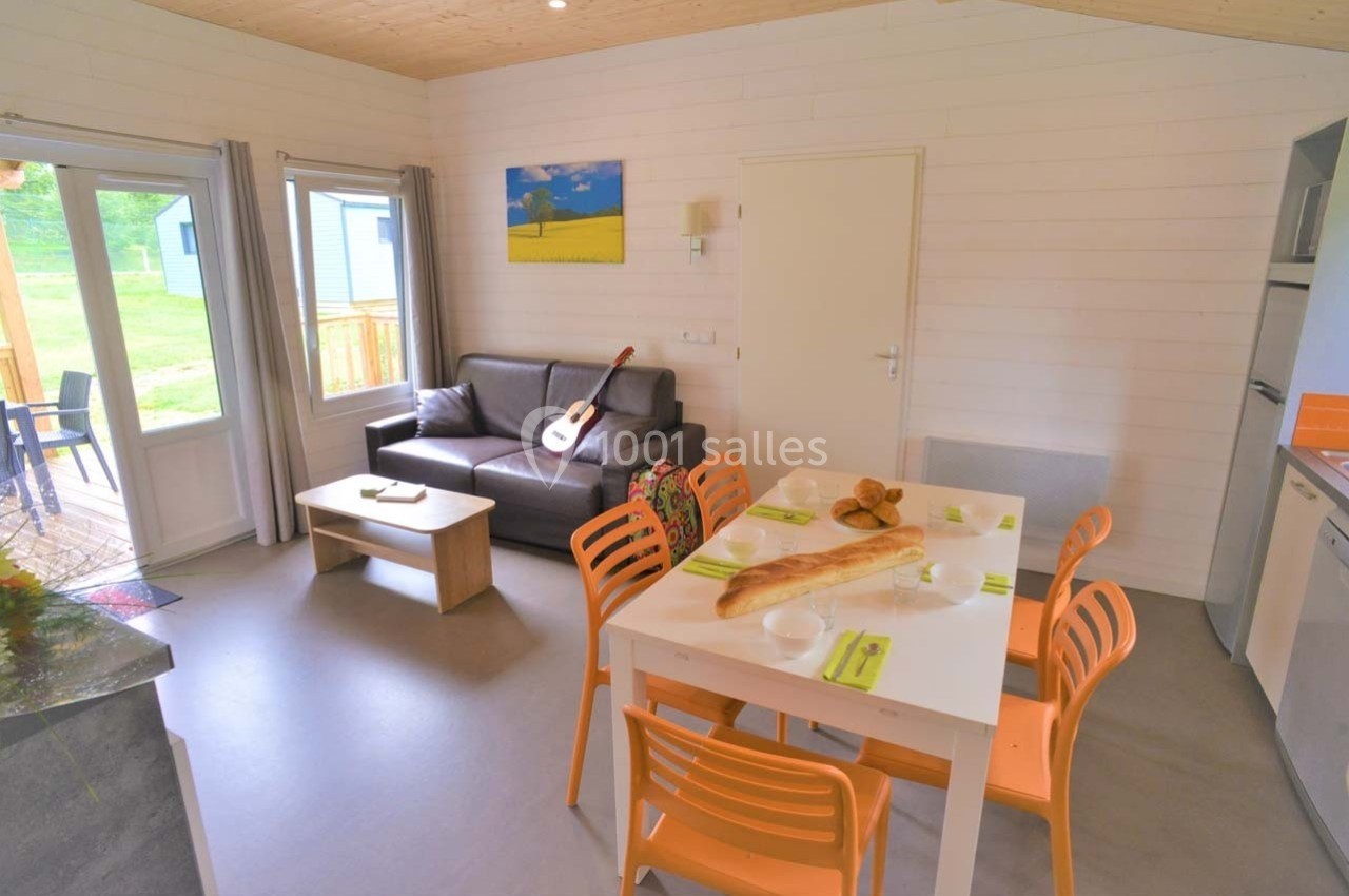 Location salle Merdrignac (Côtes-d'Armor) - Val de Landrouet #18 Salon lumineux avec canapé, table à manger, chaises orange, guitare posée, et vue sur terrasse et jardin.