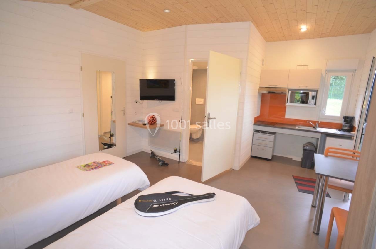 Location salle Merdrignac (Côtes-d'Armor) - Val de Landrouet #21 Intérieur d'un chalet avec deux lits simples, une kitchenette équipée, une table et une télévision fixée au mur.