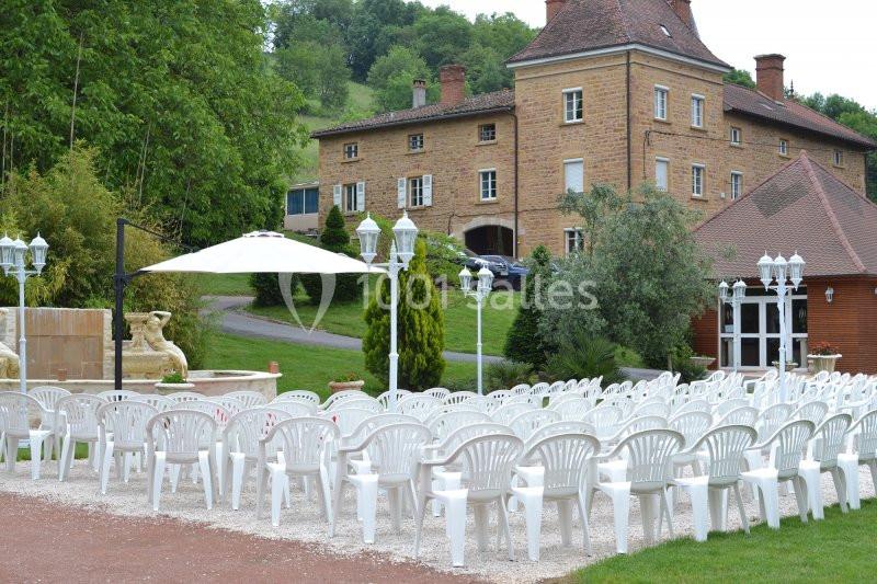 Chaises blanches disposées en rangées en extérieur devant un bâtiment en briques, entouré de verdure.