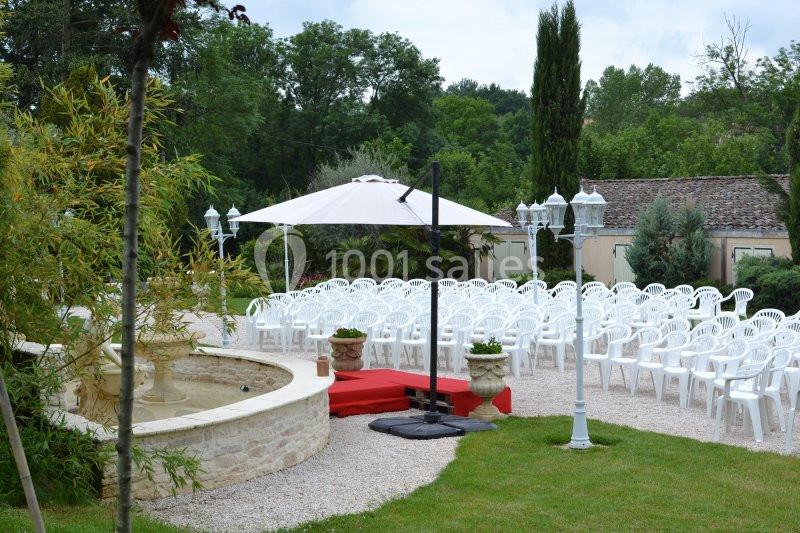 Chaises blanches disposées en extérieur devant une fontaine, avec un parasol et des lampadaires, entourés de verdure.