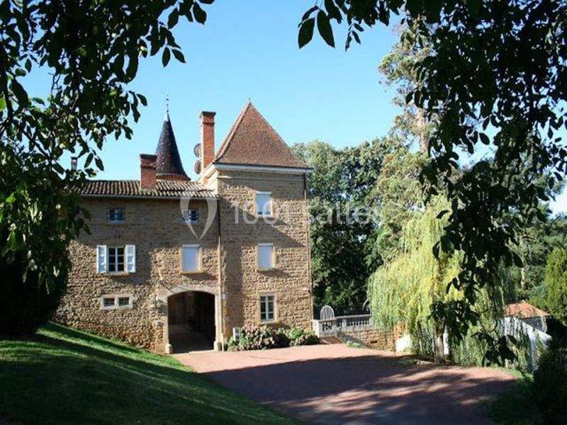 Manoir en pierre entouré de verdure, avec une allée pavée et des arbres encadrant la vue.
