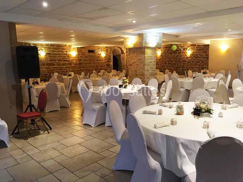 Salle de réception avec des tables rondes dressées, nappes blanches et chaises houssées, éclairage chaleureux.
