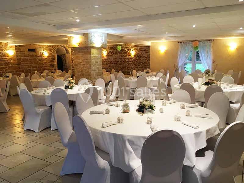Salle de réception décorée avec des tables rondes dressées, nappes blanches et chaises houssées, murs en pierre apparente.