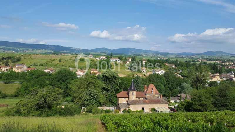 Vue d'un village entouré de verdure avec des collines et un ciel légèrement nuageux en arrière-plan.