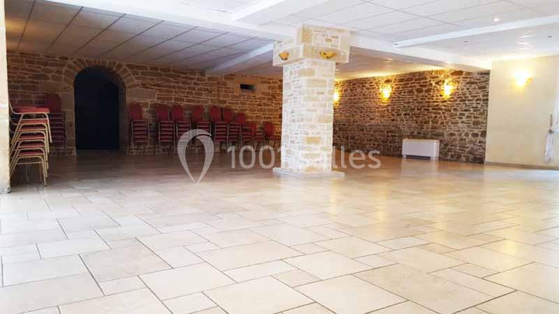 Salle spacieuse avec murs en pierre, sol carrelé clair, colonnes centrales et chaises empilées dans un coin.