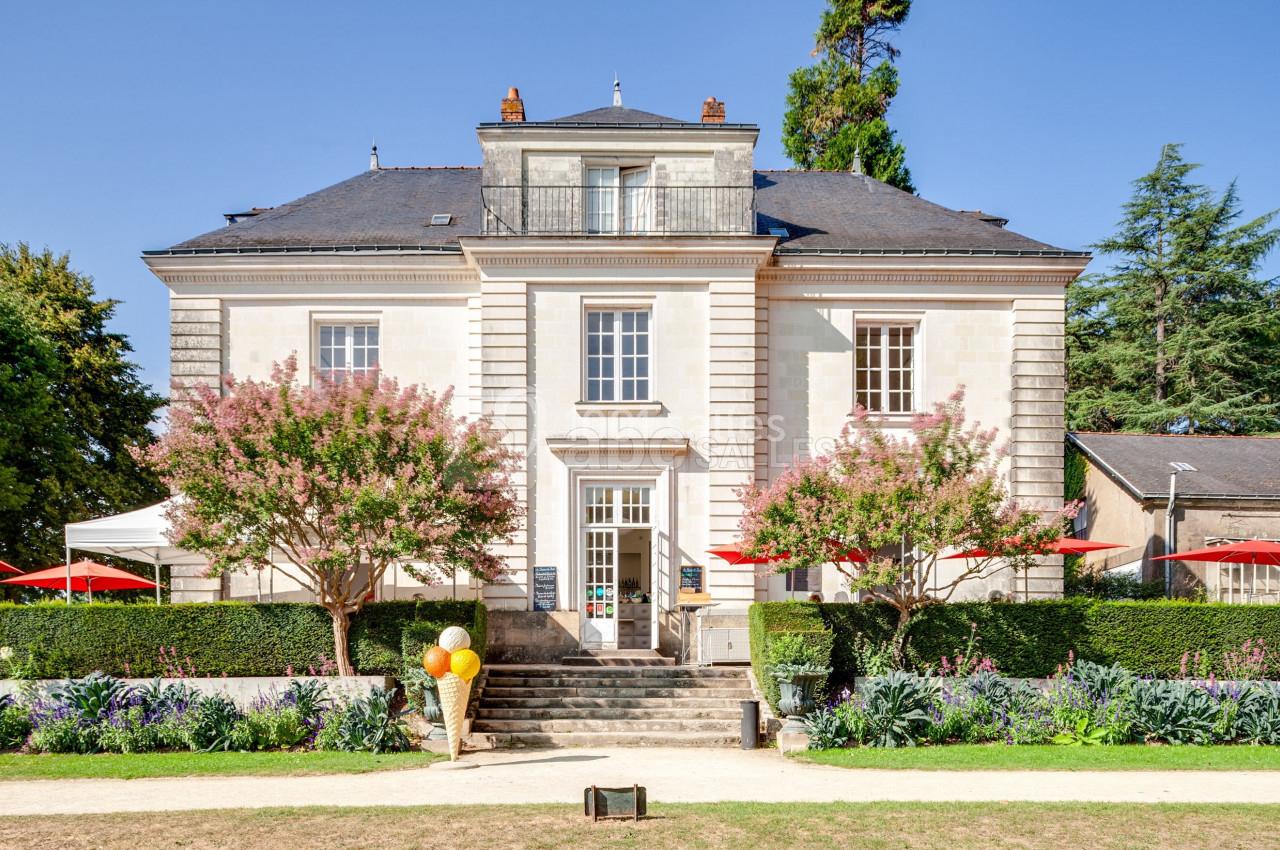 Façade d'une maison bourgeoise entourée de jardins, avec des arbres en fleurs et des parasols rouges.