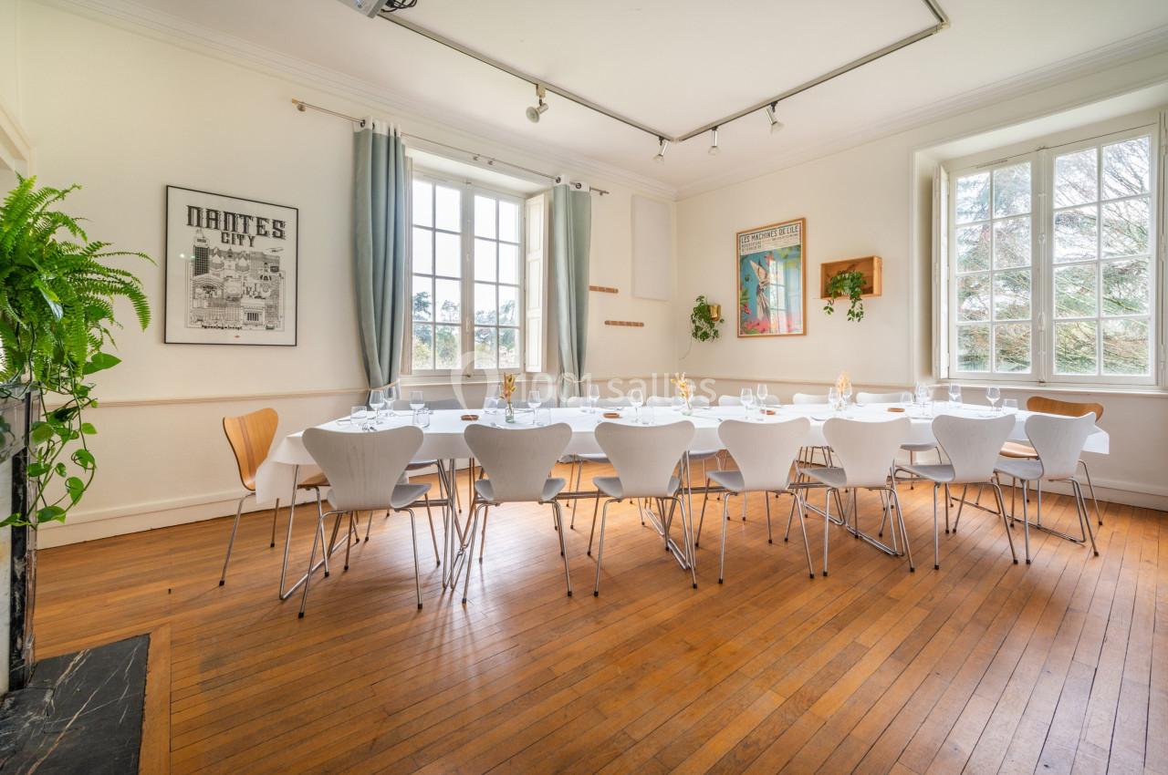 Salle lumineuse avec grande table dressée, chaises modernes, parquet en bois et fenêtres donnant sur un extérieur verdoyant.