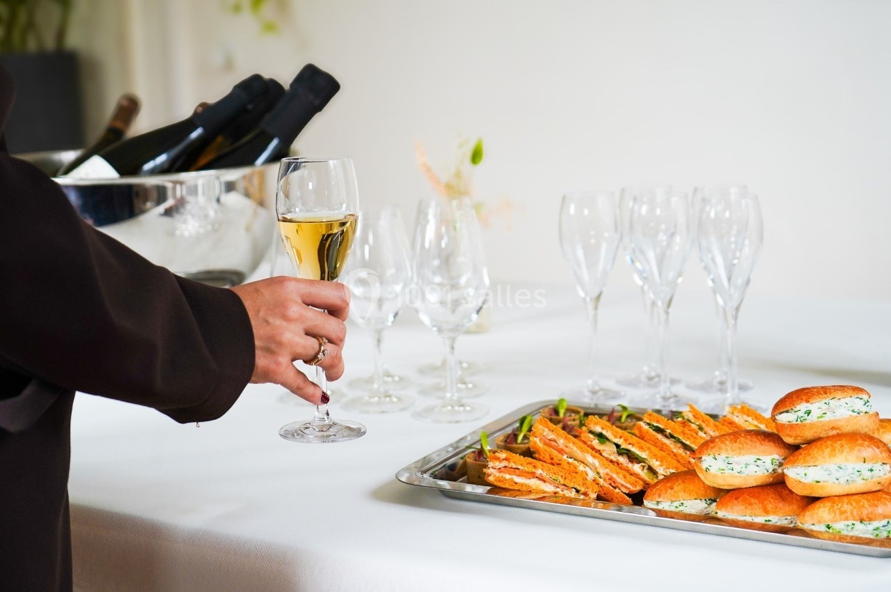 Une main tenant un verre de vin blanc près d'une table avec des verres vides, des bouteilles et des sandwichs.