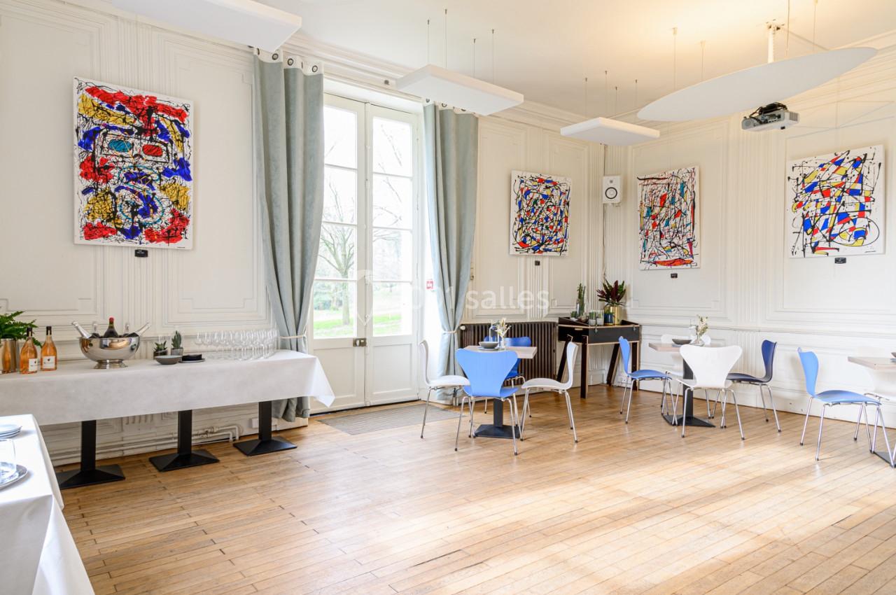 Salle lumineuse avec parquet, tables dressées, chaises bleues, grandes fenêtres et tableaux colorés accrochés aux murs.