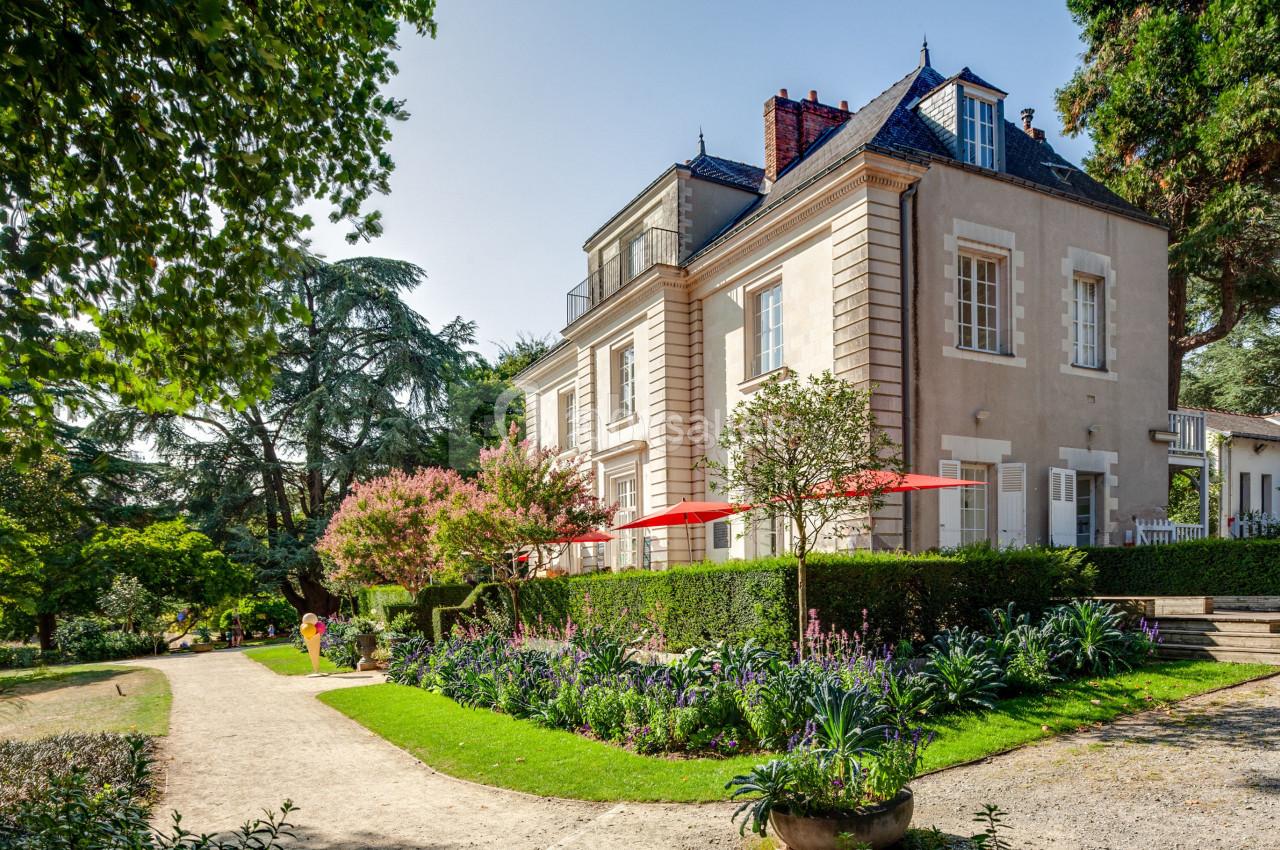 Grande maison en pierre entourée d'un jardin fleuri avec allée, arbres et parasols rouges sur la terrasse.