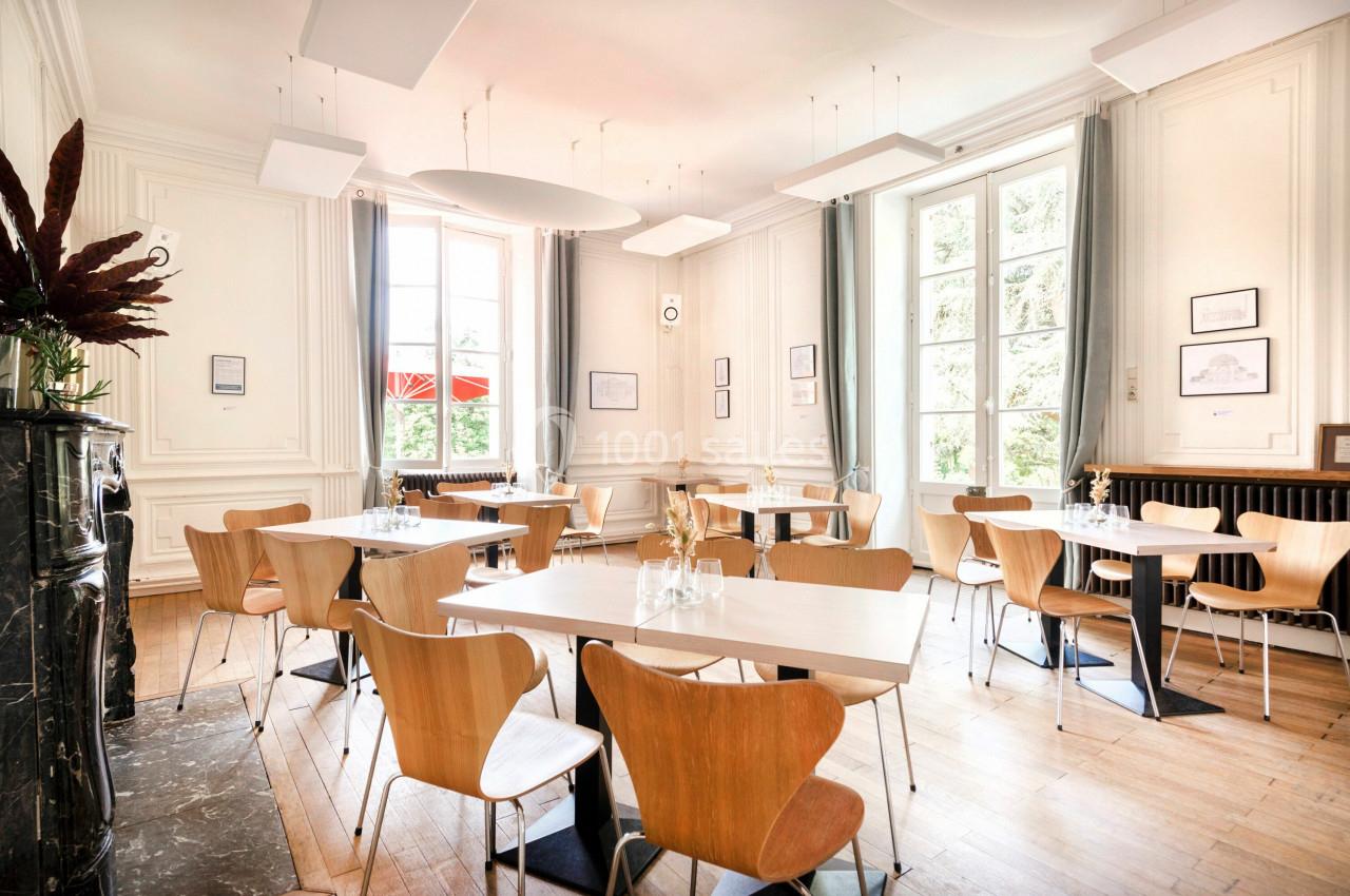 Salle lumineuse avec des tables et des chaises en bois, grandes fenêtres, murs blancs et décoration minimaliste.