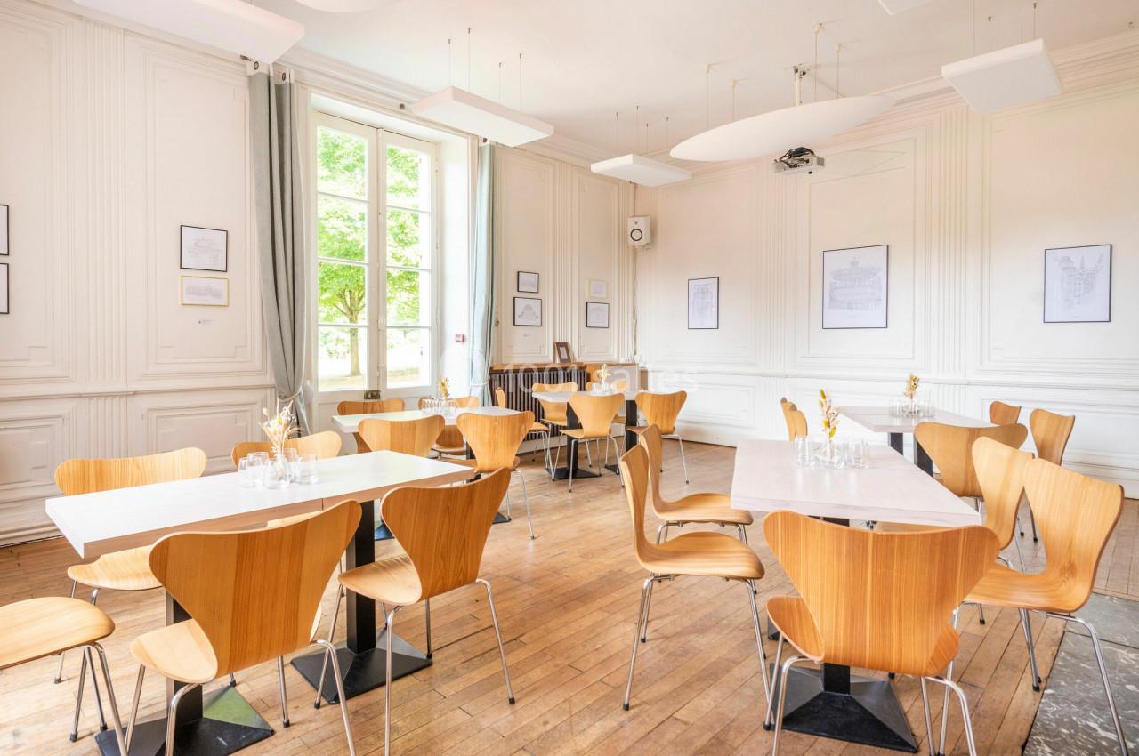 Salle lumineuse avec parquet, tables blanches, chaises en bois clair et grandes fenêtres donnant sur un extérieur verdoyant.