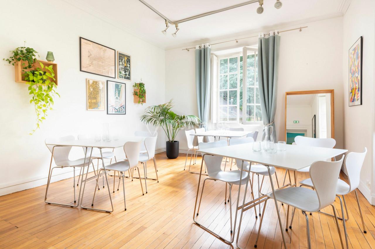 Salle lumineuse avec parquet, tables blanches, chaises modernes, plantes, cadres au mur et grande fenêtre avec rideaux.