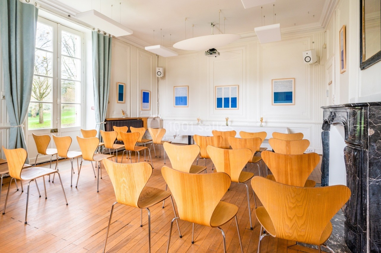 Salle lumineuse avec des chaises en bois disposées face à un écran, parquet au sol et grandes fenêtres donnant sur un jardin.